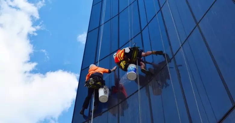 dois alpinistas industriais içados em uma fachada de vidro executando a limpeza de prédios