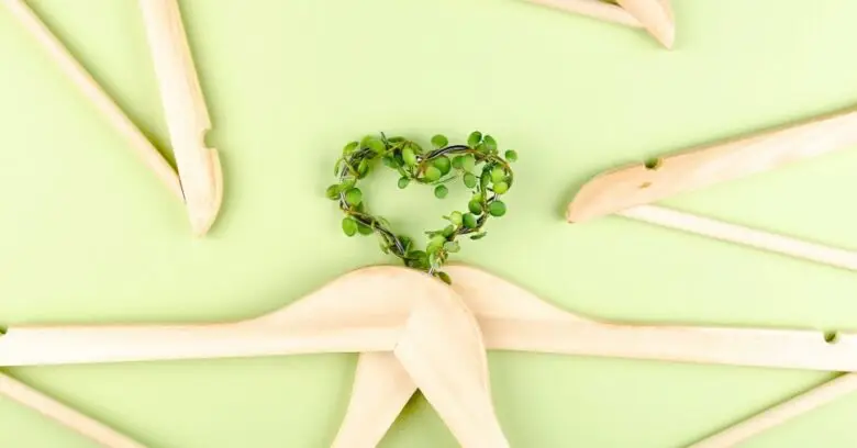 Cabides de madeira e coração de folhas sobre fundo verde ilustram práticas sustentáveis na moda.